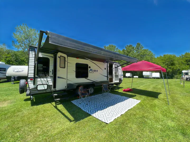 Sunny RV rental setup: travel trailer with awning, outdoor rug, chairs, and red tent on a grassy campsite.. Palomino Puma XLE 25 2018