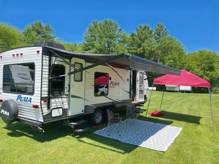 Puma travel trailer RV rental setup on green grass with an extended awning, red pop-up canopy, outdoor rug, and chairs for a relaxing camp trip.. Palomino Puma XLE 25 2018