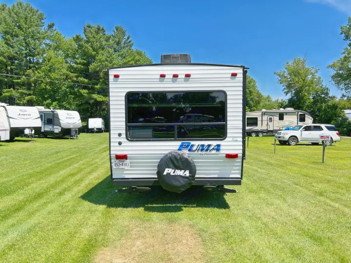 Rear of a white Puma travel trailer for RV rental, parked in a sunny grassy field with blue sky and other RVs.. Palomino Puma XLE 25 2018