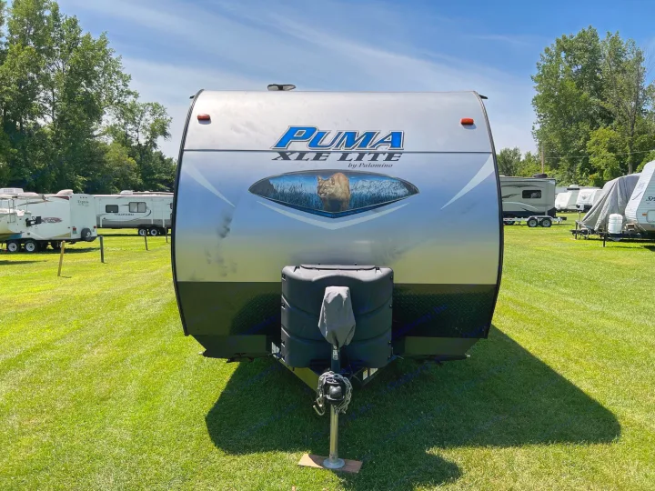 Front of a silver Puma XLE Lite travel trailer, ideal for RV rental, parked on green grass with other campers under sunny skies.. Palomino Puma XLE 25 2018