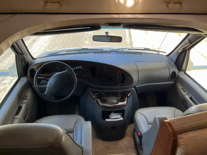 Inside an RV rental's front cabin, showing the driver's area, dashboard, and passenger seats from a rear perspective.. Itasca Sundancer 2001