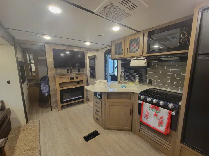 Bright RV rental interior with a modern kitchen featuring a sink and stove, a living area with TV/fireplace, and light wood flooring.. Keystone RV Hideout 2021