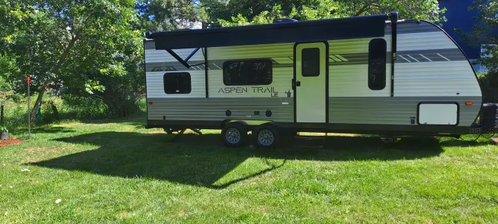 RV rental: A white and gray travel trailer with an extended awning is parked on green grass among trees, ready for camping.. Dutchmen Other 2022