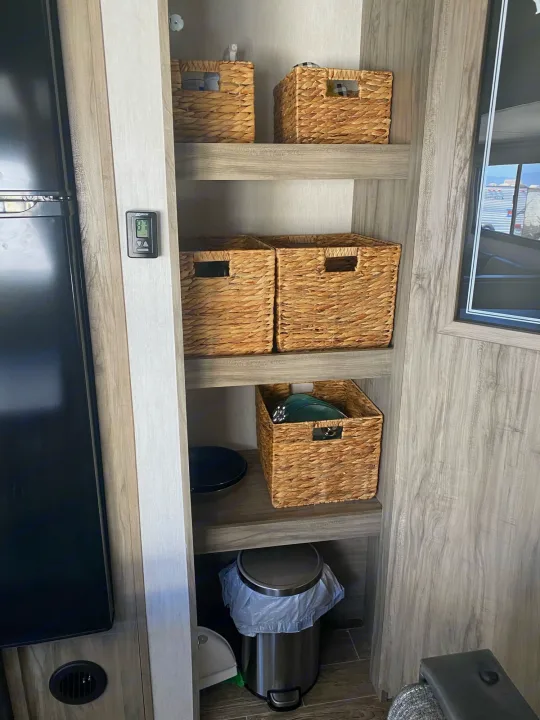 Inside an RV rental: a wood shelving unit with woven baskets, a thermostat, and a trash can for organized living.. Forest River Cherokee 2022