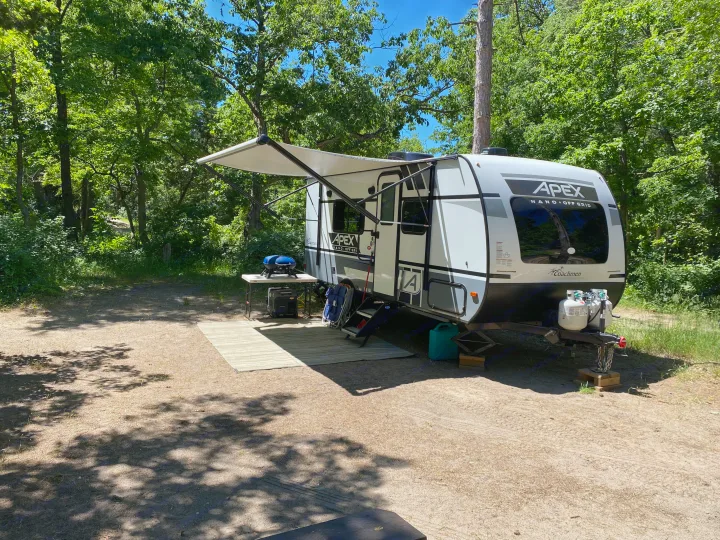 White RV rental with awning extended, grill, and camping gear, parked on a dirt lot under green trees at a sunny campsite.. Coachmen Apex 2021