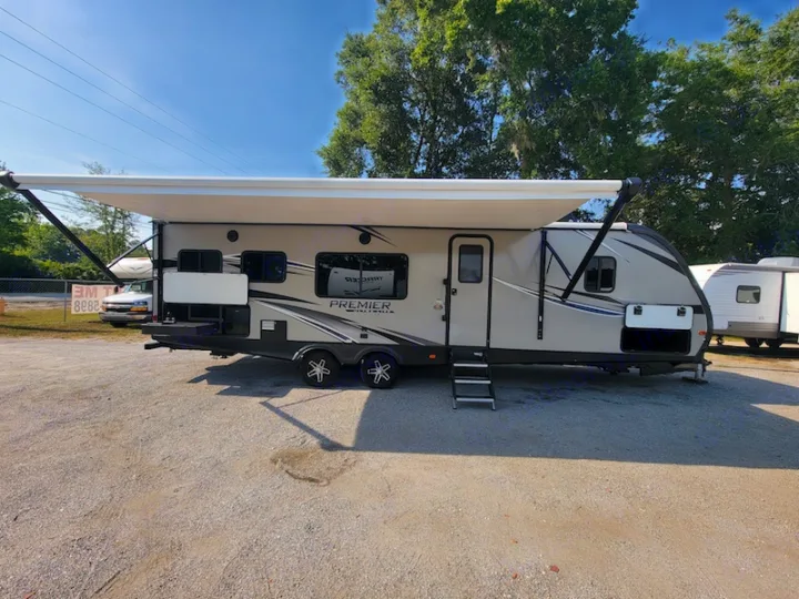 Light-colored PREMIER travel trailer RV rental parked on gravel with awning extended on a sunny day.. Keystone RV Premier UltraLite 29RKPR 2021