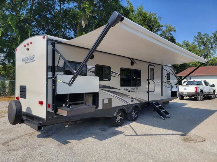 Spacious RV rental travel trailer with an extended awning and outdoor kitchen setup on a gravel lot, ready for adventure.. Keystone RV Premier UltraLite 29RKPR 2021