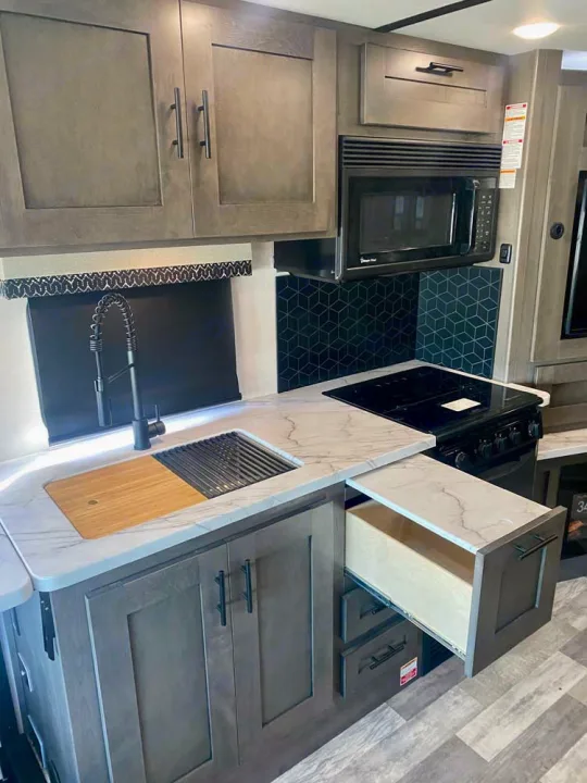 Modern RV rental kitchen with grey cabinets, white marbled countertops, sink with accessories, stove, and microwave.. Forest River FR3 34DL 2020