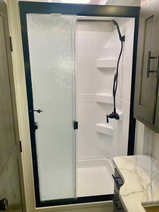 Inside an RV rental: a modern, compact shower with frosted glass sliding door, black fixtures, and a white stall.. Forest River FR3 34DL 2020
