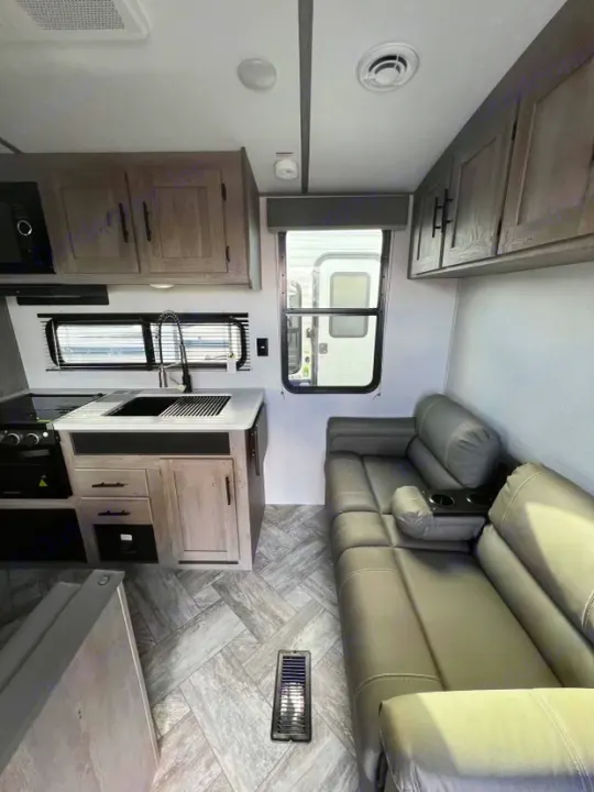 Modern RV rental interior: light wood cabinetry kitchen with sink and stove, plus a comfy grey sectional sofa on herringbone flooring.. Keystone RV Springdale 2021