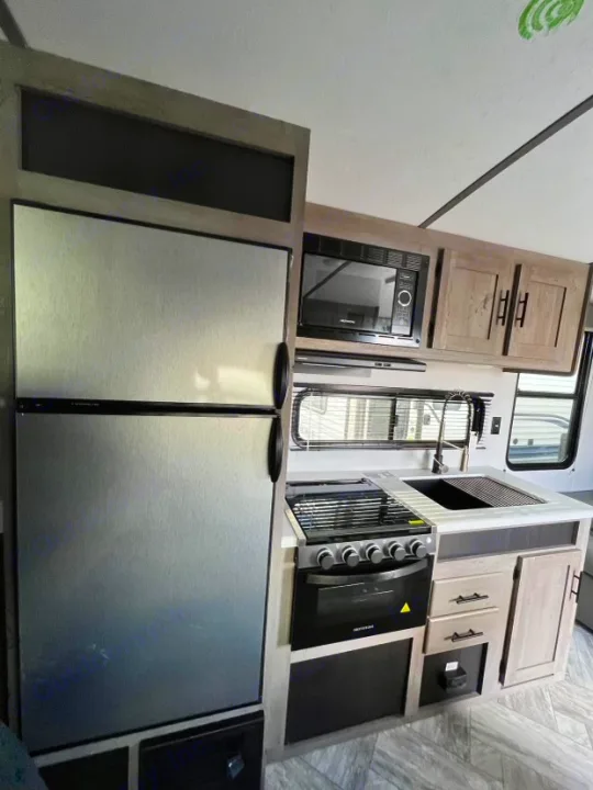 Inside an RV rental: a compact kitchen featuring a stainless steel fridge, stove, oven, sink, microwave, and wood cabinets.. Keystone RV Springdale 2021
