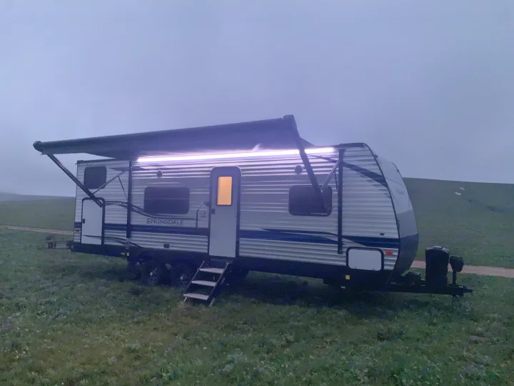 A bright, illuminated RV rental with an extended awning parked in a grassy field at twilight.. Keystone RV Springdale 2021