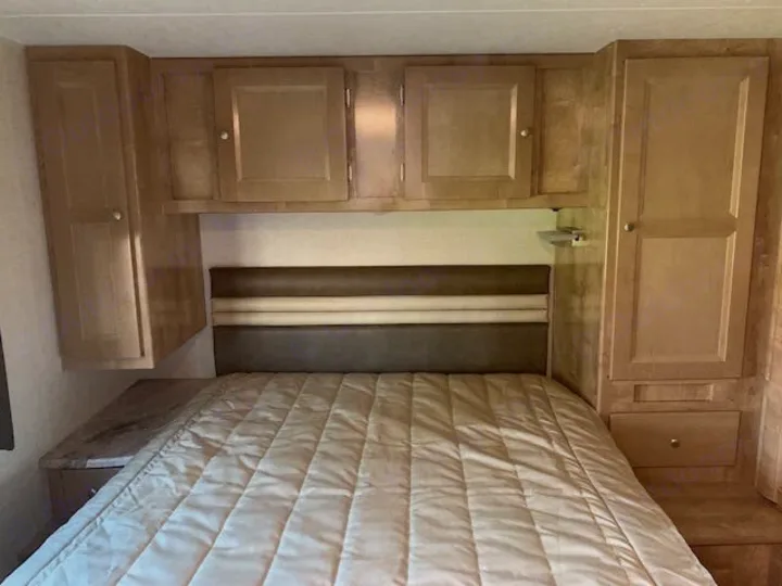 Cozy RV rental bedroom with ample cabinet storage above and beside the bed, plus an upholstered headboard.. Forest River Coachmen Leprechaun 2019