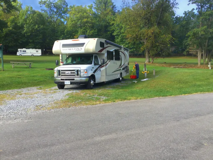 This RV rental is parked at a grassy campsite. A person is connecting hookups, ideal for a relaxing outdoor travel experience.. Forest River Coachmen Leprechaun 2019