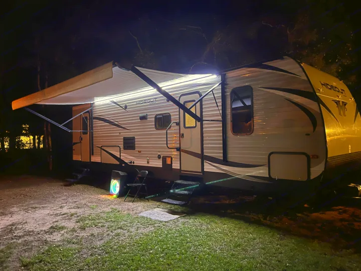An RV rental parked at night, brightly lit by its awning LEDs, steps, and a colorful speaker, offering a cozy outdoor space.. Keystone RV Hideout 2018