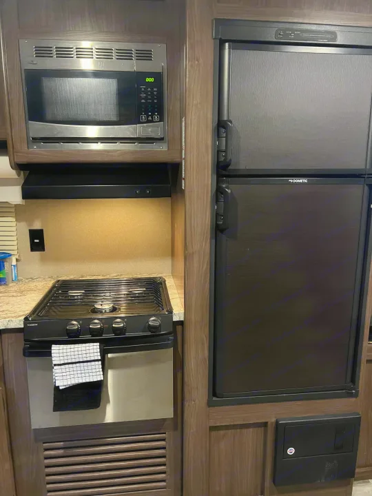 An RV rental kitchen showcasing a stainless steel microwave, black gas stove with oven, and two-door Dometic refrigerator.. Keystone RV Hideout 2018