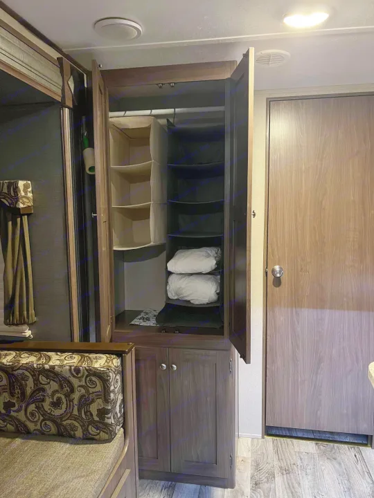 RV rental interior: open wardrobe with hanging storage, folded items, and built-in lower cabinets next to textured cushions and a closed door.. Keystone RV Hideout 2018