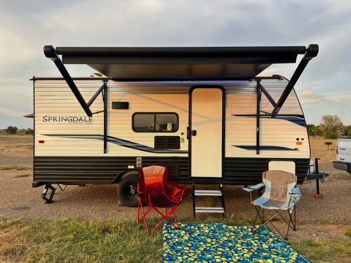 Springdale travel trailer RV rental parked at a serene campsite with awning, chairs, and mat ready for leisure.. Keystone Springdale 2022