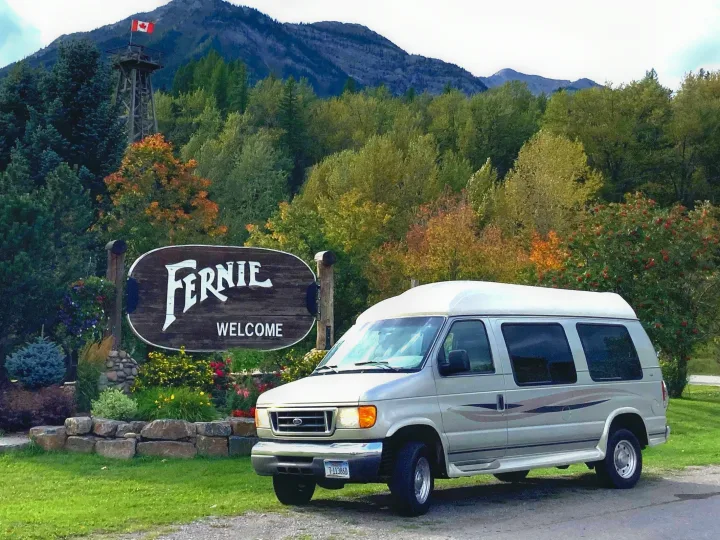 Silver RV rental van at Fernie, BC welcome sign, with Canadian flag, mountains & autumn trees.. Ford Econoline 2006