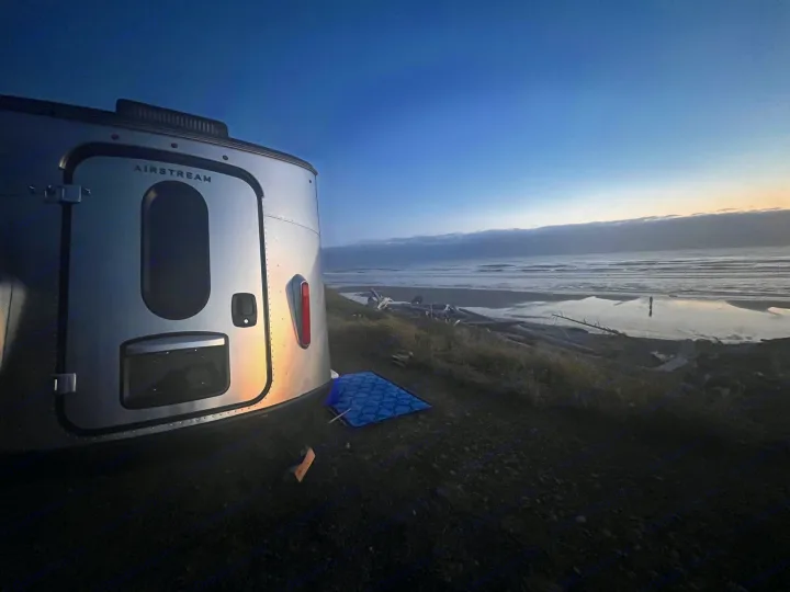 An Airstream rv rental overlooks a tranquil beach at dusk. The silver travel trailer sits by the ocean's edge.. Airstream Basecamp 2022