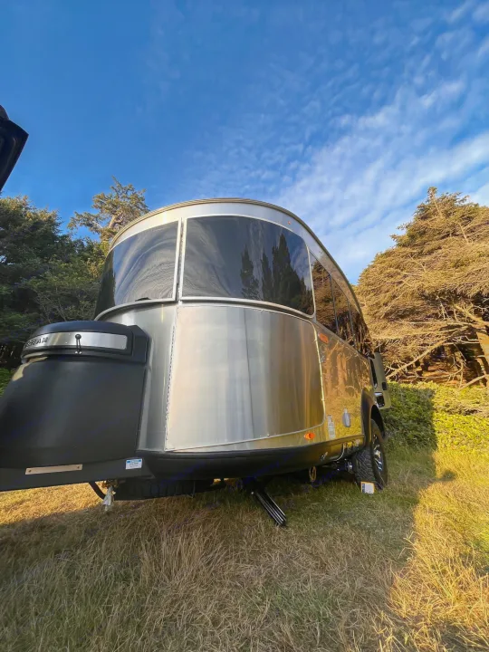 Reflective Airstream travel trailer set up as an rv rental under a blue sky, surrounded by trees.. Airstream Basecamp 2022