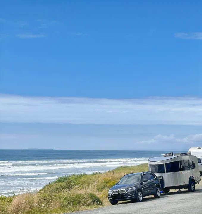 A black SUV & a silver rv rental parked on a coastal road, with ocean waves and a distant island under a blue sky.. Airstream Basecamp 2022