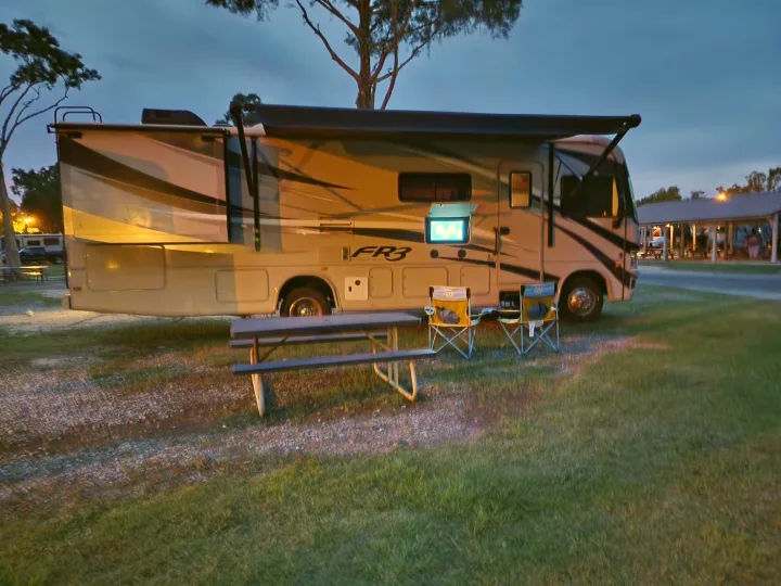 An RV rental provides comfort at dusk: a motorhome with an outdoor TV, chairs, and picnic table on a grassy site.. Forest River Fr3 2015
