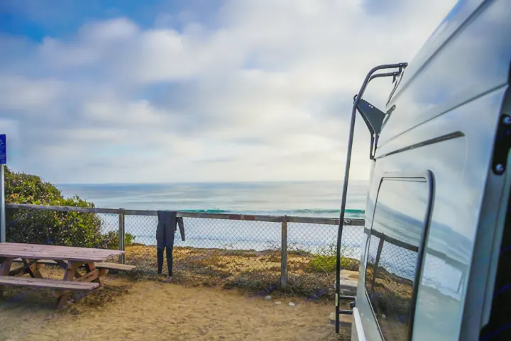 RV rental parked on a sandy bluff, overlooking the ocean with waves & surfers. A wetsuit hangs on the fence near a picnic table.. Winnebago Revel 2022