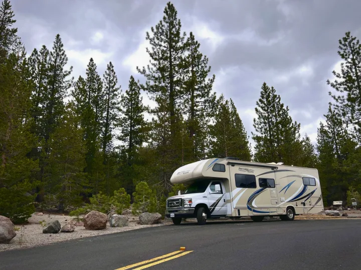 A Class C RV rental parked on a road surrounded by dense pine trees and rocks under an overcast sky.. Thor Freedom Elite 2019