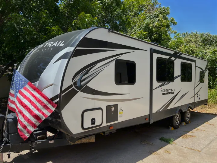 A white North Trail Ultra Lite travel trailer with American flag attached, parked outdoors. Ideal RV rental.. Heartland RVs North Trail 2021