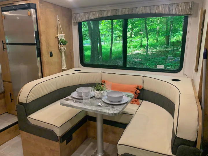 This RV rental's dinette area features a U-shaped booth with a set table, fridge, and large window overlooking a forest.. Forest River Coachmen Leprechaun 2020