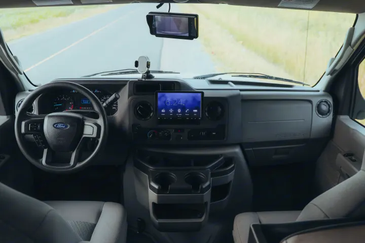 Interior of an RV rental van: dashboard, steering wheel, and a digital screen showing 6:24 PM. Road visible through windshield.. Forester LE 2021