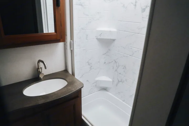 Bright RV rental bathroom with a white marble-look shower-tub, corner shelves, and a modern sink with a dark counter.. Forester LE 2021