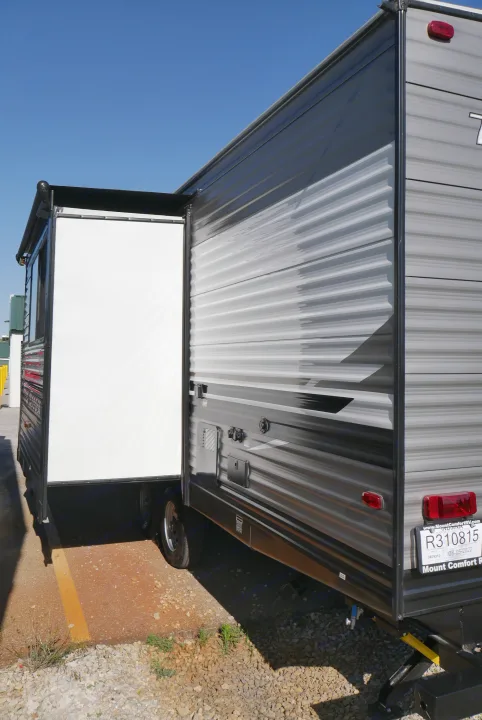 This modern RV rental opens its side to reveal an outdoor entertainment screen, set on a gravel lot under blue sky.. Heartland RVs Trailrunner 2021