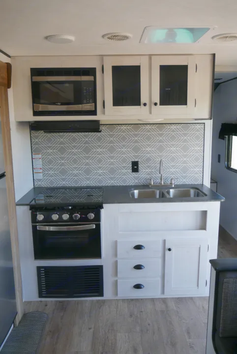 Inside an RV rental: a well-equipped kitchen with a stove, microwave, double sink, and white storage cabinets.. Heartland RVs Trailrunner 2021