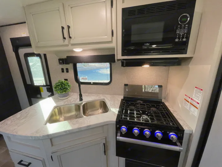 Modern RV rental kitchen with double sink, gas range (blue knob lights), microwave, and light cabinets.. Jayco Jay Flight 2022