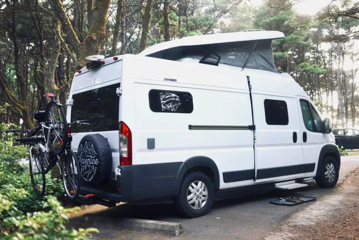 White Hymer RV rental in a peaceful forest, featuring bikes, a pop-top roof, and outdoor shoes for nature travel.. Hymer Hymer Class B 2019