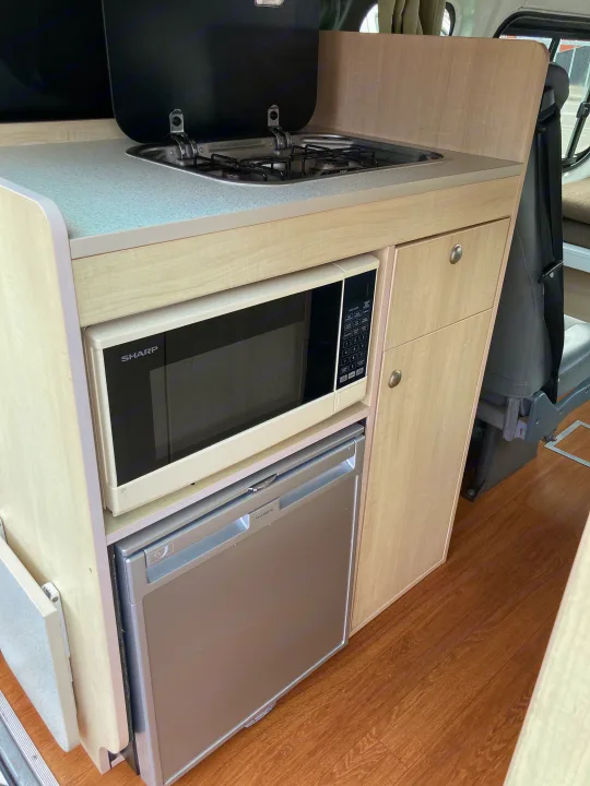 RV rental kitchenette featuring a built-in stovetop, Sharp microwave, and Dometic refrigerator with light wood finishes.. Toyota Hiace 2013