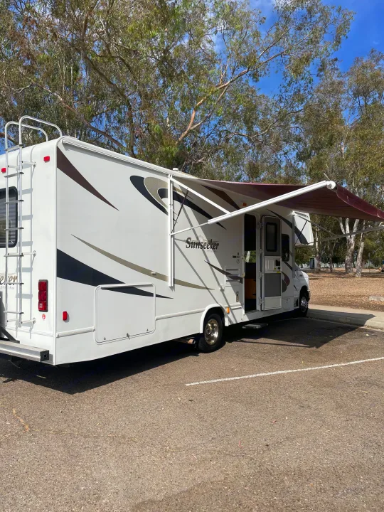 RV rental: A white Sunseeker RV with an extended awning is parked in an outdoor lot under trees, ready for use.. Forest River Sunseeker 2009