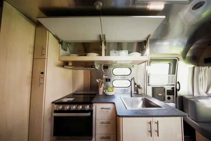 Spacious modern rv rental kitchen featuring a cooktop, oven, sink, and open cabinets with stainless steel walls.. Airstream Flying Cloud 2021