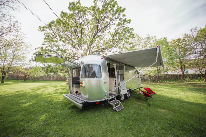 A sleek silver Airstream rv rental on a green lawn, featuring an open back hatch, extended awning, and red chairs.. Airstream Flying Cloud 2021