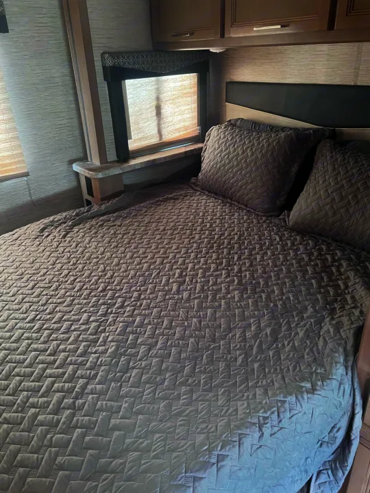 RV rental interior showing a comfortable bedroom with a grey quilted bedspread, headboard, and window.. Thor Motor Coach Four Winds 2021