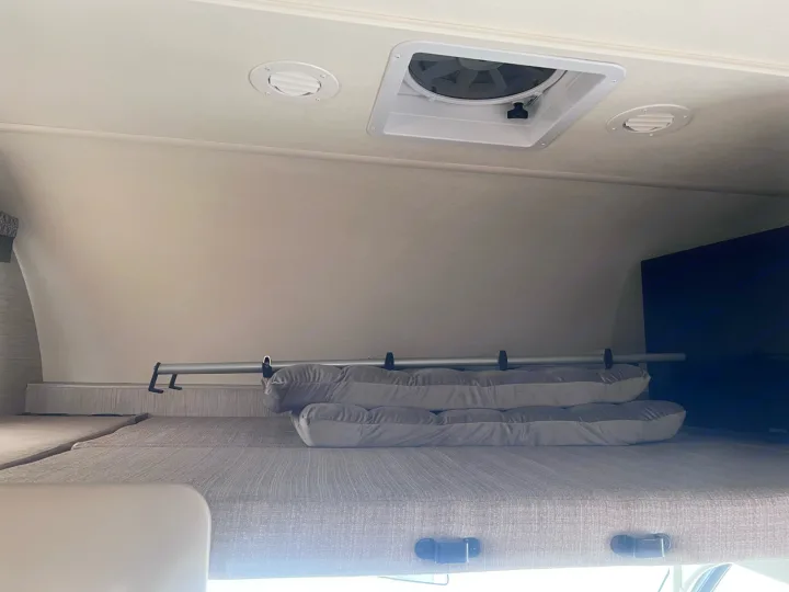 Inside an RV rental: overhead bunk bed with safety rail, stacked cushions, ceiling vent, and TV screen on the right.. Thor Motor Coach Four Winds 2021