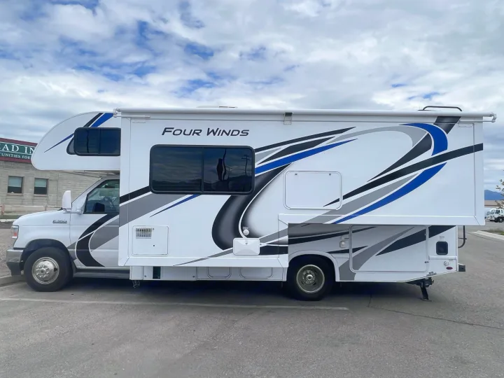 This white Four Winds Class C RV rental, with blue and gray graphics, is parked on asphalt under a cloudy sky.. Thor Motor Coach Four Winds 2021