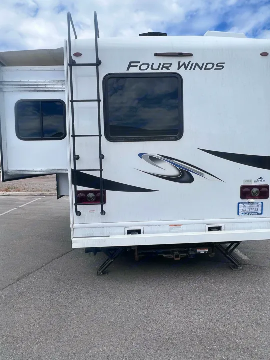 Rear of a white Four Winds RV rental, showcasing an extended slide-out, roof ladder, and stabilizing jacks for your next road trip.. Thor Motor Coach Four Winds 2021