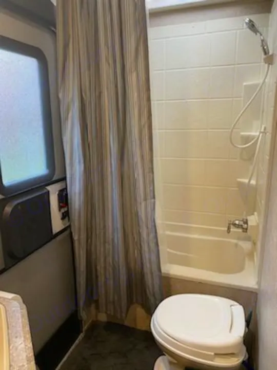 Inside an RV rental bathroom, showing a white toilet, compact bathtub with shower, and a gray striped shower curtain.. Forest River Coachmen Catalina SBX 2017