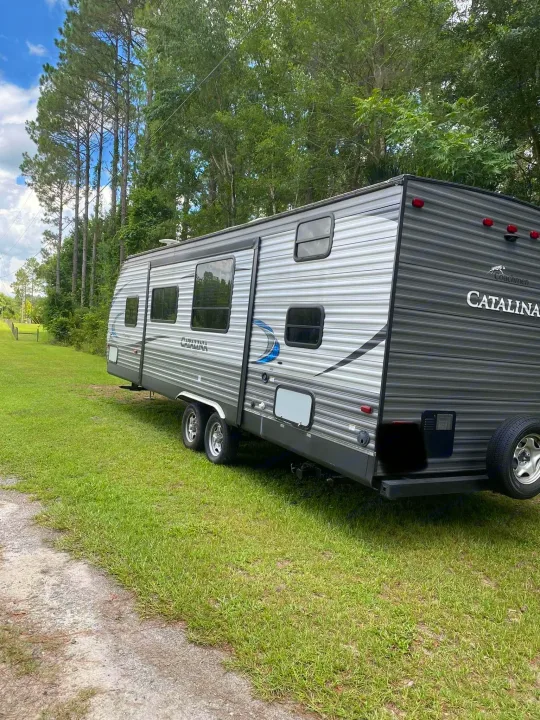 Gray Coachmen Catalina RV rental, a sleek travel trailer, parked on green grass next to a path, surrounded by tall trees.. Forest River Coachmen Catalina SBX 2017