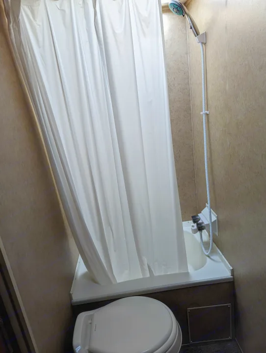 View of a compact RV rental bathroom, including a white shower curtain, small tub, handheld shower, and toilet.. Forest River Cherokee Grey Wolf 2015