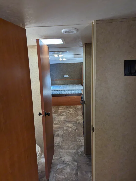 RV rental interior, showing a hallway leading to a bedroom with a bed. A bathroom door on the left reveals a toilet.. Forest River Cherokee Grey Wolf 2015