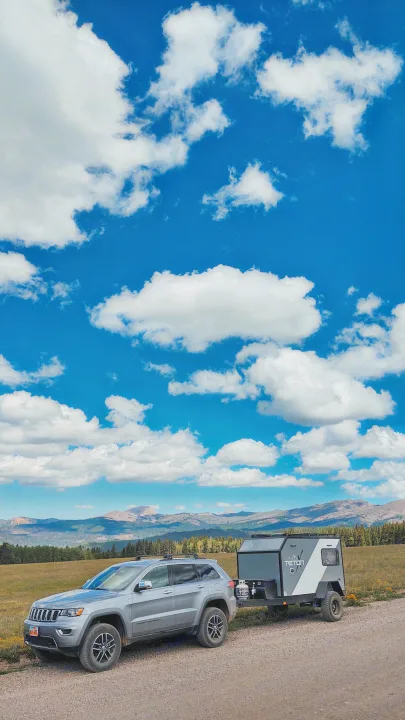 Silver SUV towing a Teton C camper trailer, an ideal RV rental for mountain adventures, parked on a dirt road with scenic mountains & blue sky.. TetonX Porter 2022